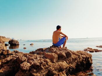 Man sitting on rock at beach against clear sky