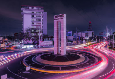 Light trails on road by buildings against sky at night