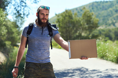 Portrait of young man holding cardboard while standing on road against sky