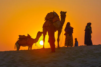 Silhouette of people and camels on desert against orange sunset sky