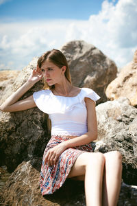 Young woman sitting on rock against sky