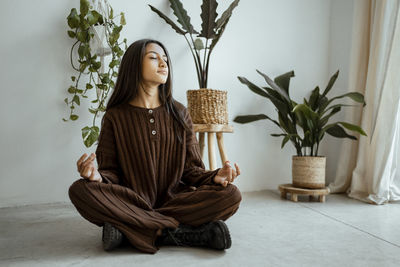 Beautiful girl with eyes closed sitting in lotus position against potted plants at home
