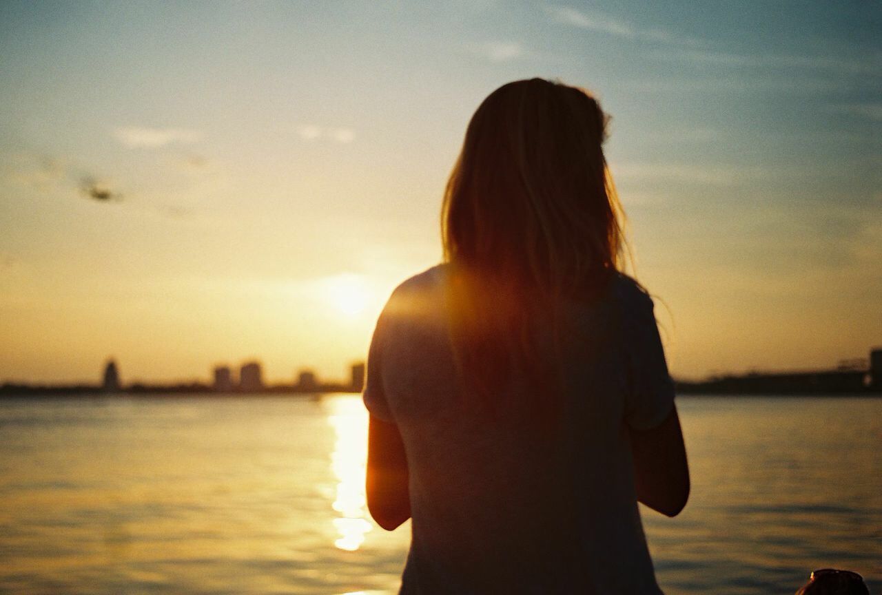 water, sunset, rear view, lifestyles, sea, waist up, sky, leisure activity, standing, three quarter length, person, silhouette, focus on foreground, tranquility, side view, long hair, looking at view, nature
