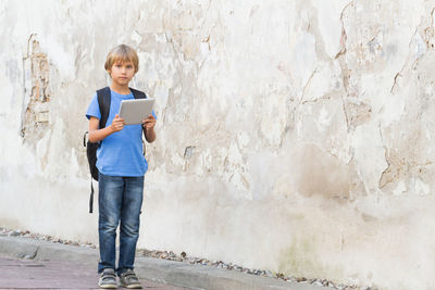 Full length portrait of cute boy holding digital tablet while standing by wall