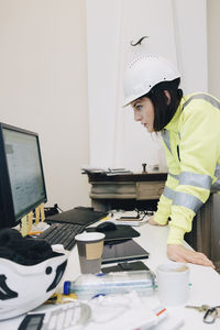 Female building contractor in hardhat using computer while leaning on desk at office