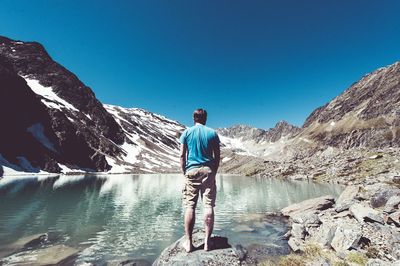 Rear view of man standing on rock by lake against clear blue sky