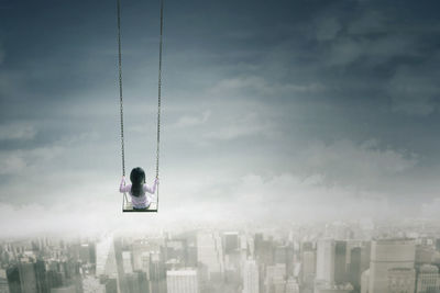 Digital composite image of girl swinging over buildings against sky