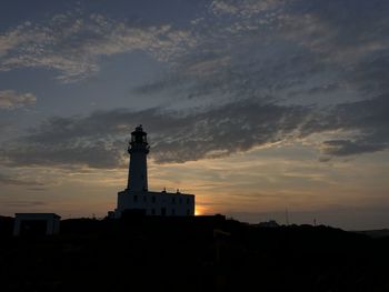 Silhouette lighthouse by building against sky during sunset
