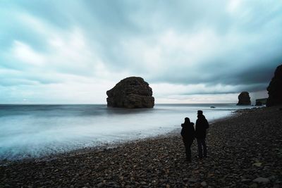 People standing on pebbles stones at beach against sky