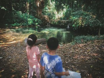 Rear view of siblings standing by lake in forest