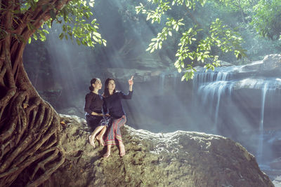 Friends sitting on rock against waterfall in forest