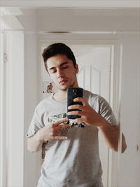 Young man taking selfie with smart phone reflecting on mirror at home