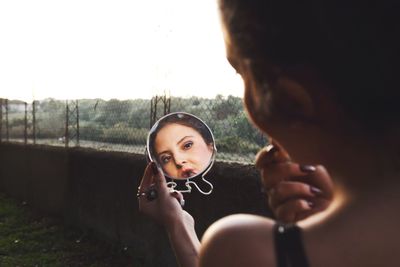 Rear view of young woman applying lipstick reflecting on mirror while standing against clear sky