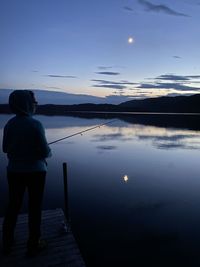 Rear view of man standing by lake against sky during sunset