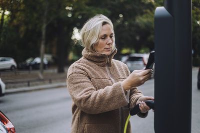 Woman holding charger while scanning through smart phone at charging station