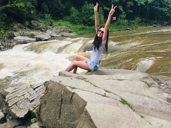 Portrait of smiling woman with arms raised sitting on rock at riverbank