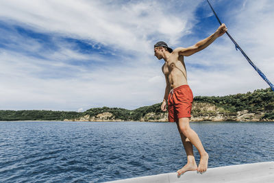 Shirtless man looking at sea while standing on boat against cloudy sky during sunny day