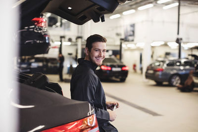 Portrait of happy young mechanic leaning on car trunk