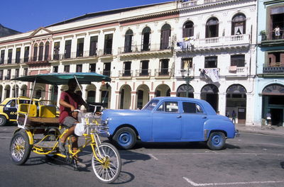 Man riding pedicab by vintage car on street against building