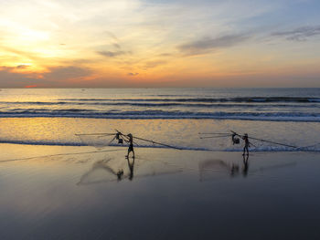 Fishermen with fishing nets standing at beach against sky during sunset