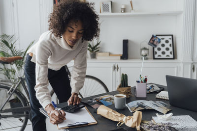 Freelancer standing at hert desk, using calculater, taking notes