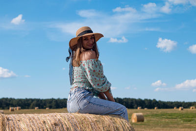 A beautiful young ukrainian woman with hat sits on a hay bale and smiles in to the camera