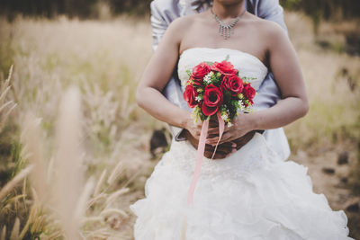 Midsection of bride and bridegroom holding bouquet on field