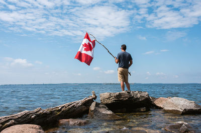 Man holding canada flag on rocky shore of a lake on a summer day.