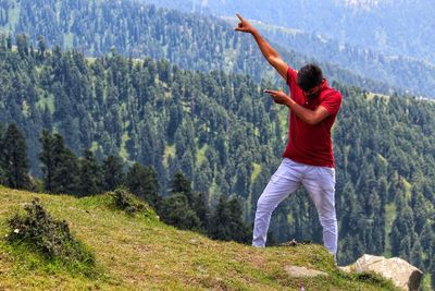 Full length of young man gesturing while standing on mountain against trees
