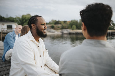 Smiling man looking away while sitting with friend on pier near lake