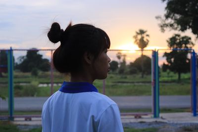 Rear view of thoughtful young woman looking away while standing at park during sunset