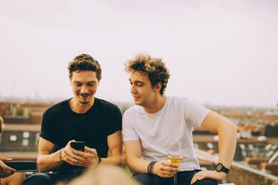 Man showing mobile phone to friend having drink at terrace during party