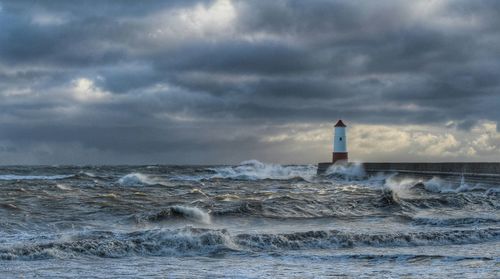 Sea waves rushing towards lighthouse against cloudy sky