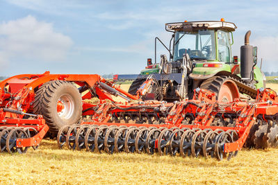 A powerful agricultural tractor combined with a red multi-row disc harrow on a summer day.