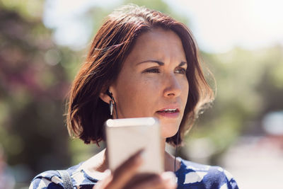 Thoughtful businesswoman looking away while holding smart phone in park during summer