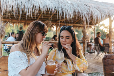 Two women sitting in beach bar at golden hour, drinking cocktails on a straw