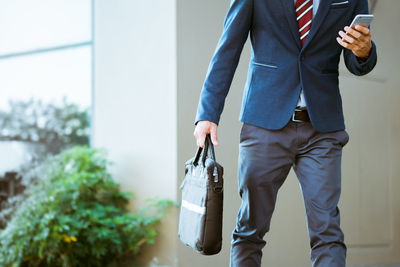 Midsection of businessman using mobile phone while walking against wall