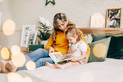 Mom and daughter are reading a book and smiling, sitting at home on the bed. new year, christmas