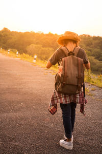 Rear view of woman with backpack standing on road against sky during sunset