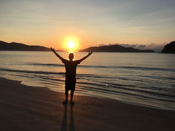 Rear view silhouette of man with arms outstretched standing at beach during sunset