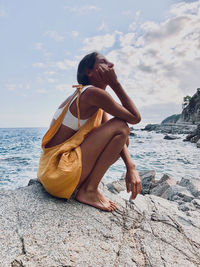 Young woman wearing bright yellow jumpsuit, sitting on a rock over the sea.summertime barefoot girl