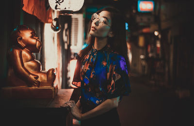 Portrait of young woman standing by statue on street at night