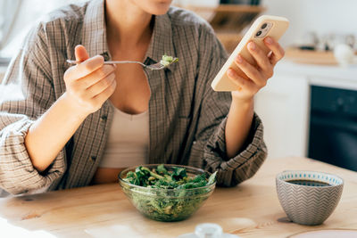 Unrecognizable modern woman having breakfast with salad and scrolling the news feed in her phone.