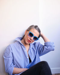 Stylish female in blue sunglasses and striped shirt touching hair while sitting on floor near wall on white background in light room