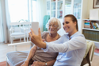 Smiling senior woman and granddaughter taking selfie using mobile phone during visit in nursing home