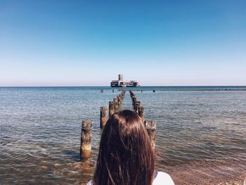 Rear view of woman standing on shore by wooden posts in sea against clear sky