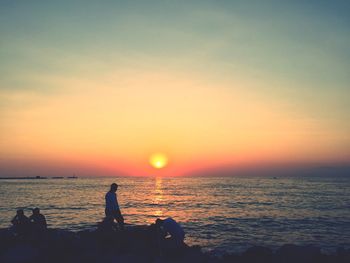 People on rocks by sea against sky during sunset