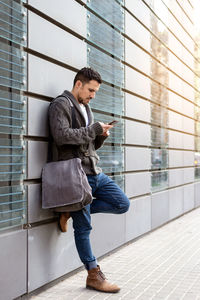 Side view of a bearded man using phone leaning on office building wall