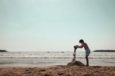 Full length of boy playing with sand while standing at beach against sky