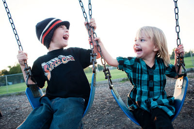 Cheerful siblings enjoying on swing against sky at playground